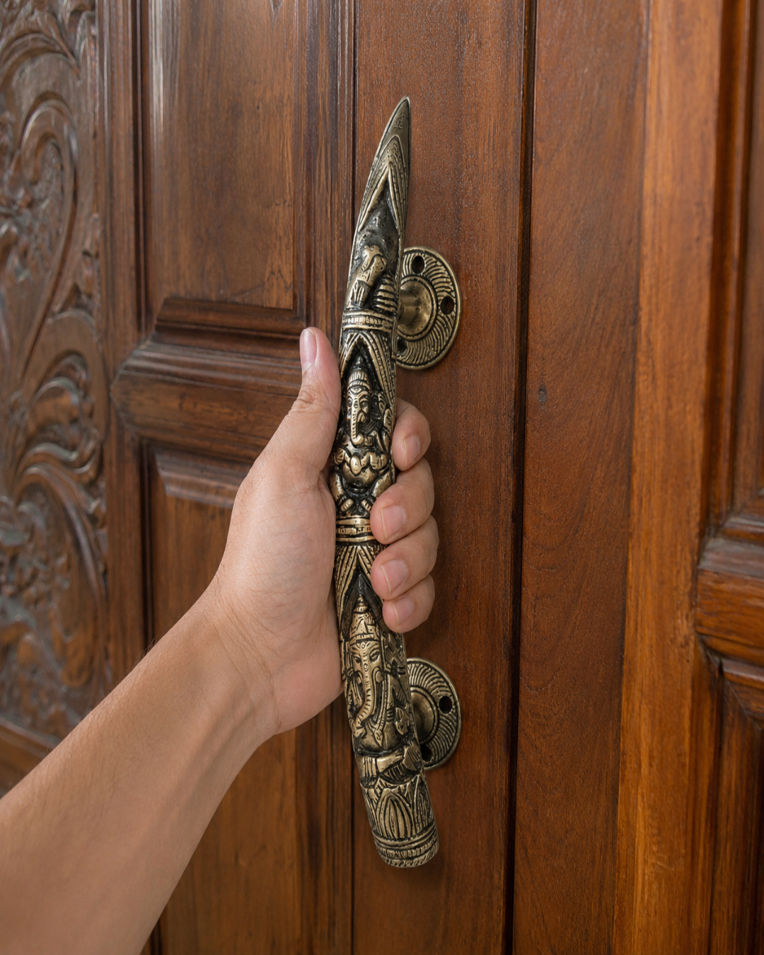 A hand holding a large, ornate brass door pull handle with detailed carvings of Lord Ganesha, set against a dark brown wooden door.