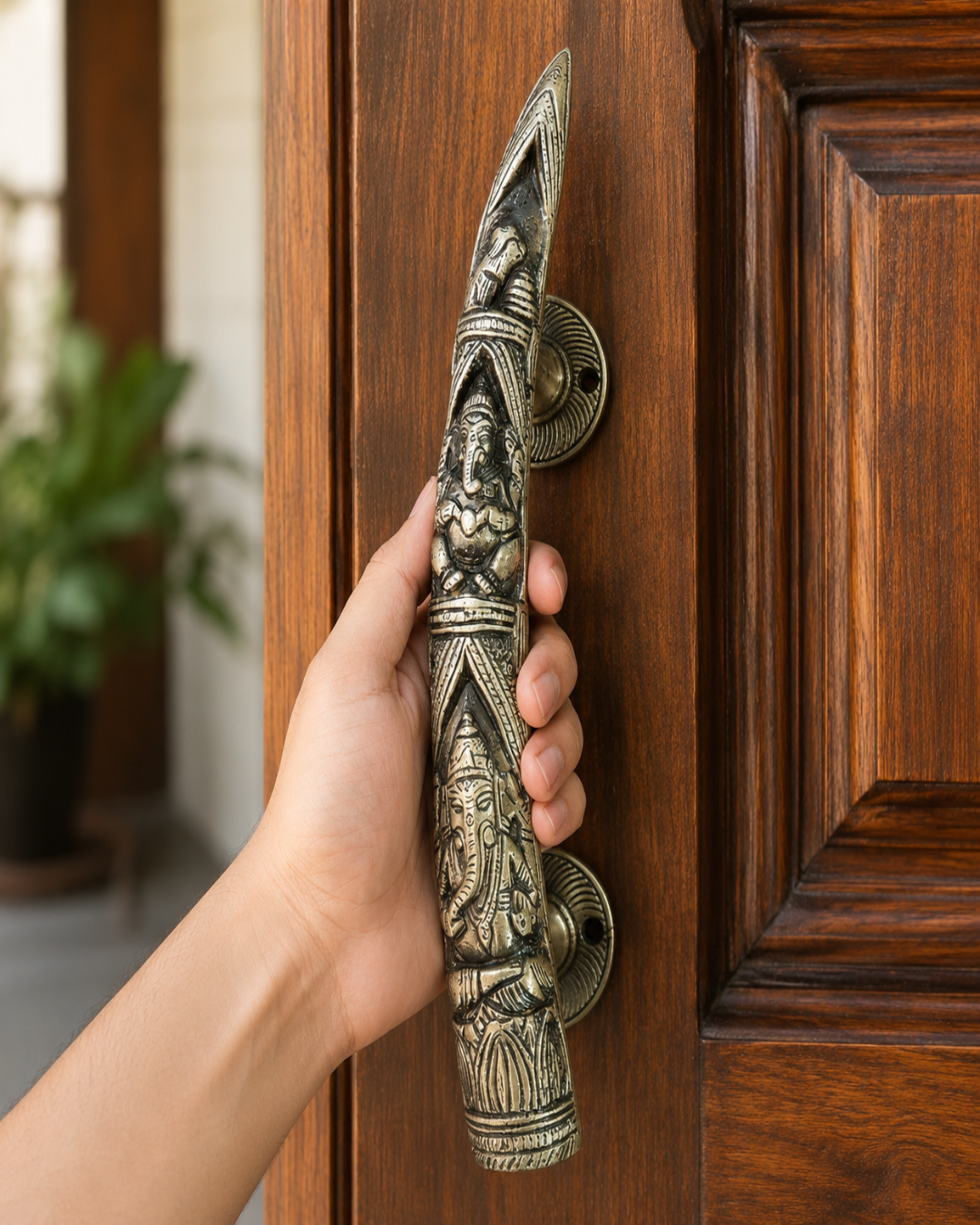 A hand holding a large, ornate bronze Ganesha door pull handle against a dark brown wooden door.