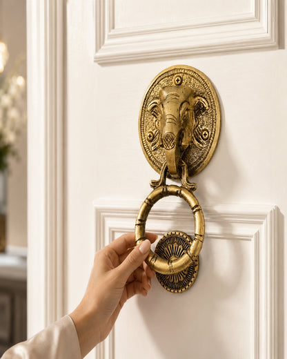 An antique-style brass elephant door knocker with a hanging ring, mounted on a white paneled door, with a hand reaching to touch it.