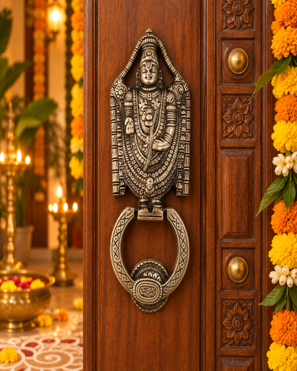 An antique-style metal door knocker featuring a detailed Hindu deity design above a circular ring, mounted on a carved dark brown wooden door decorated with marigold garlands.