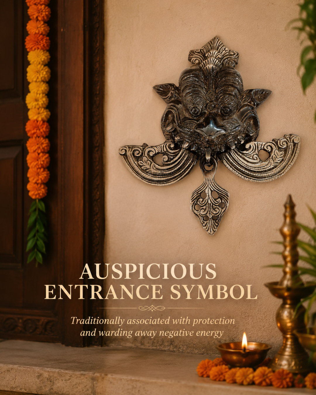An ornate metallic wall plaque depicting a traditional auspicious Ganesha symbol, mounted on a beige stucco wall next to a wooden door decorated with marigold garlands. Text overlay reads 'AUSPICIOUS ENTRANCE SYMBOL' with descriptive subtitle text below.