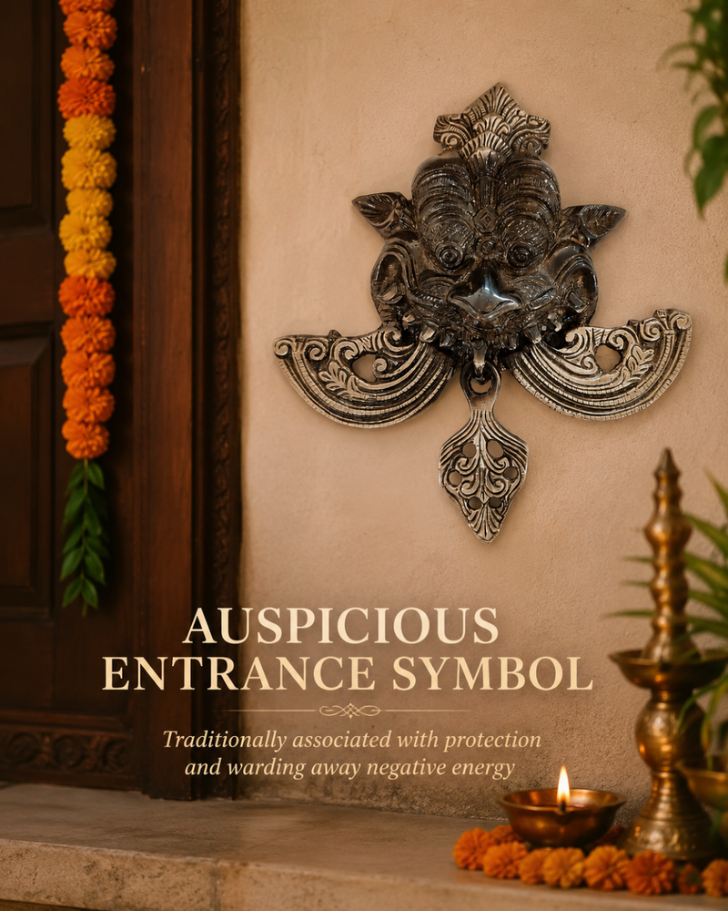 An ornate metallic wall plaque depicting a traditional auspicious Ganesha symbol, mounted on a beige stucco wall next to a wooden door decorated with marigold garlands. Text overlay reads 'AUSPICIOUS ENTRANCE SYMBOL' with descriptive subtitle text below.
