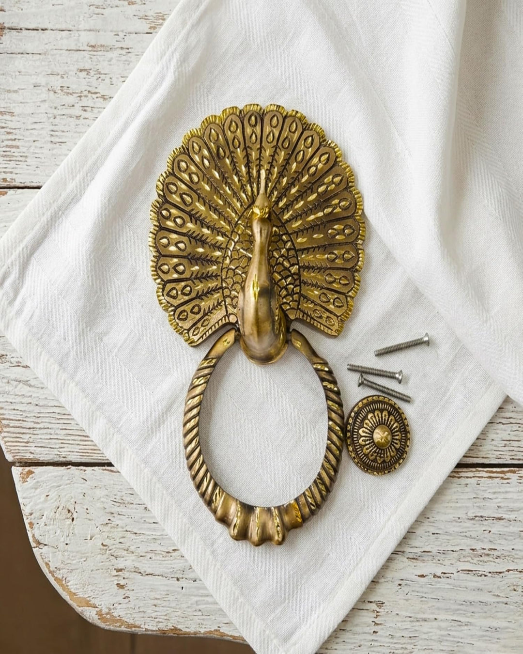 Decorative brass peacock door knocker on a white cloth with screws.