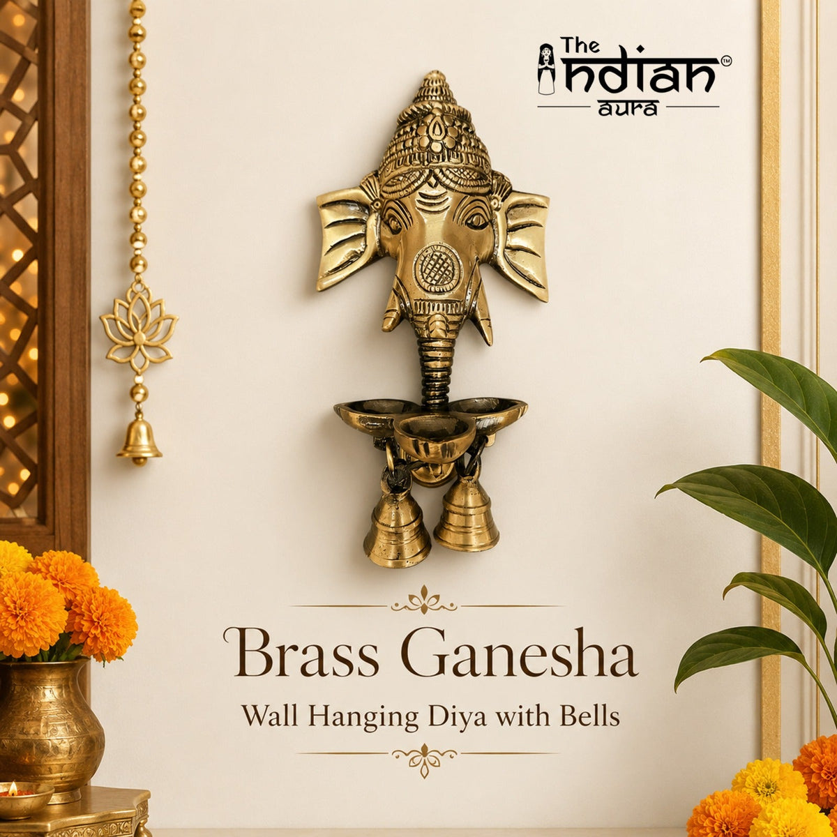A brass Ganesha wall hanging diya with bells, shown mounted on a cream wall with decorative flowers and a plant. The text 'Brass Ganesha Wall Hanging Diya with Bells' appears below, and 'The Indian Market' logo is visible at the top right.