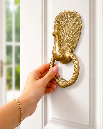 Hand using a gold peacock-shaped door knocker on a white door.