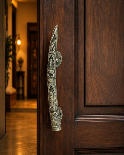 An , carved metal door handle with a traditional Indian deity design, mounted on a dark brown wooden door with a warm, softly lit hallway in the background.