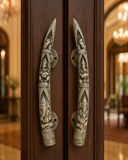 Two ornate, vertically oriented metal door handles with detailed carvings of Hindu deities, mounted on dark brown wooden doors in a warm, blurred interior setting.
