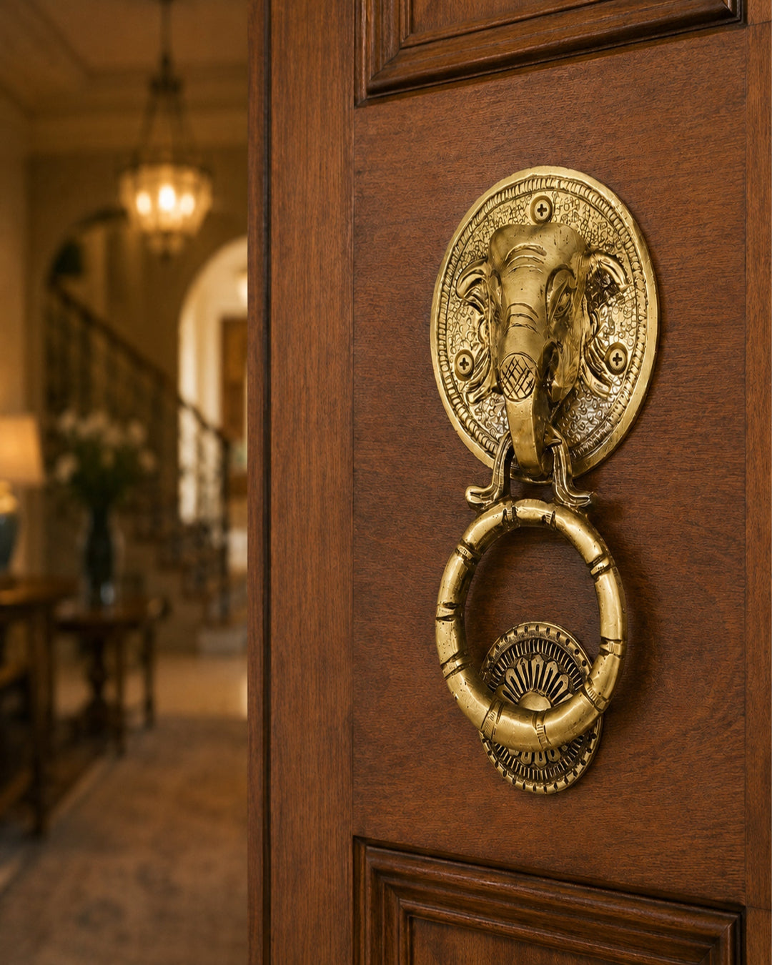 An ornate brass elephant-headed door knocker with a hanging ring, mounted on a dark brown wooden door, with a softly blurred interior hallway in the background.