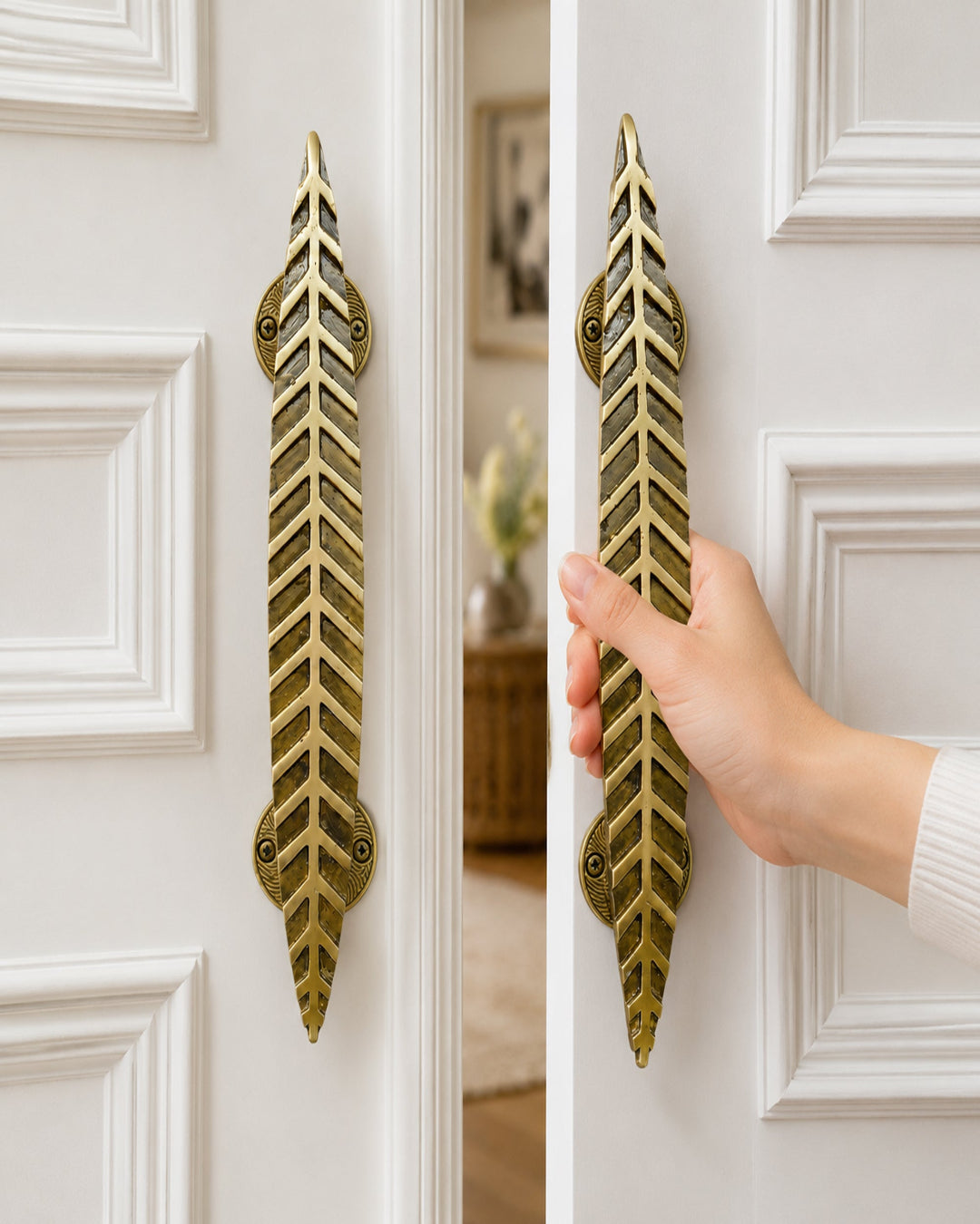 A pair of  brass door handles with a leaf-like chevron pattern, shown on white paneled doors with a hand holding the right handle open.