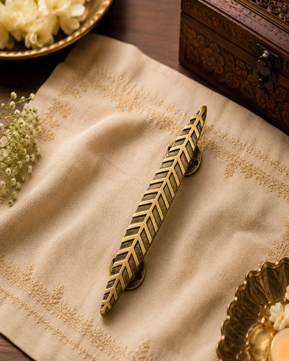 A decorative brass cabinet pull handle shaped like a leaf, displayed on a beige embroidered linen cloth with warm rustic props in the background.