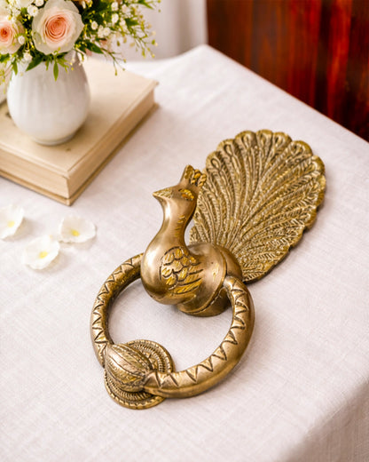 Decorative brass peacock door knocker on a white surface with flowers in the background