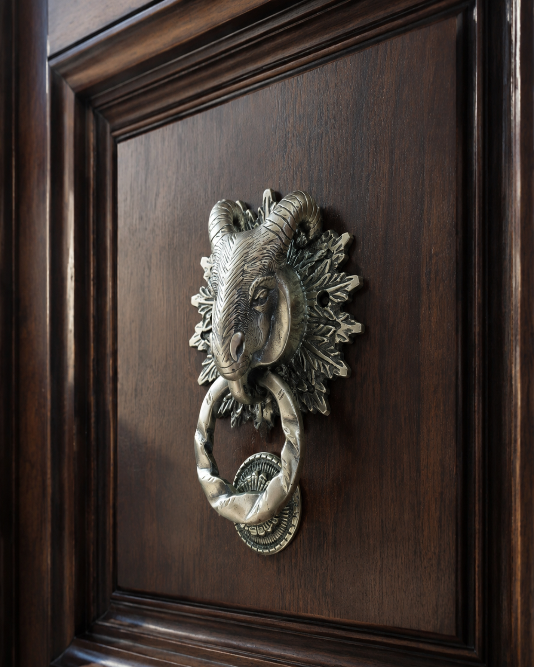 An antique-style ram head door knocker with a twisted ring, mounted on a dark brown wooden door.