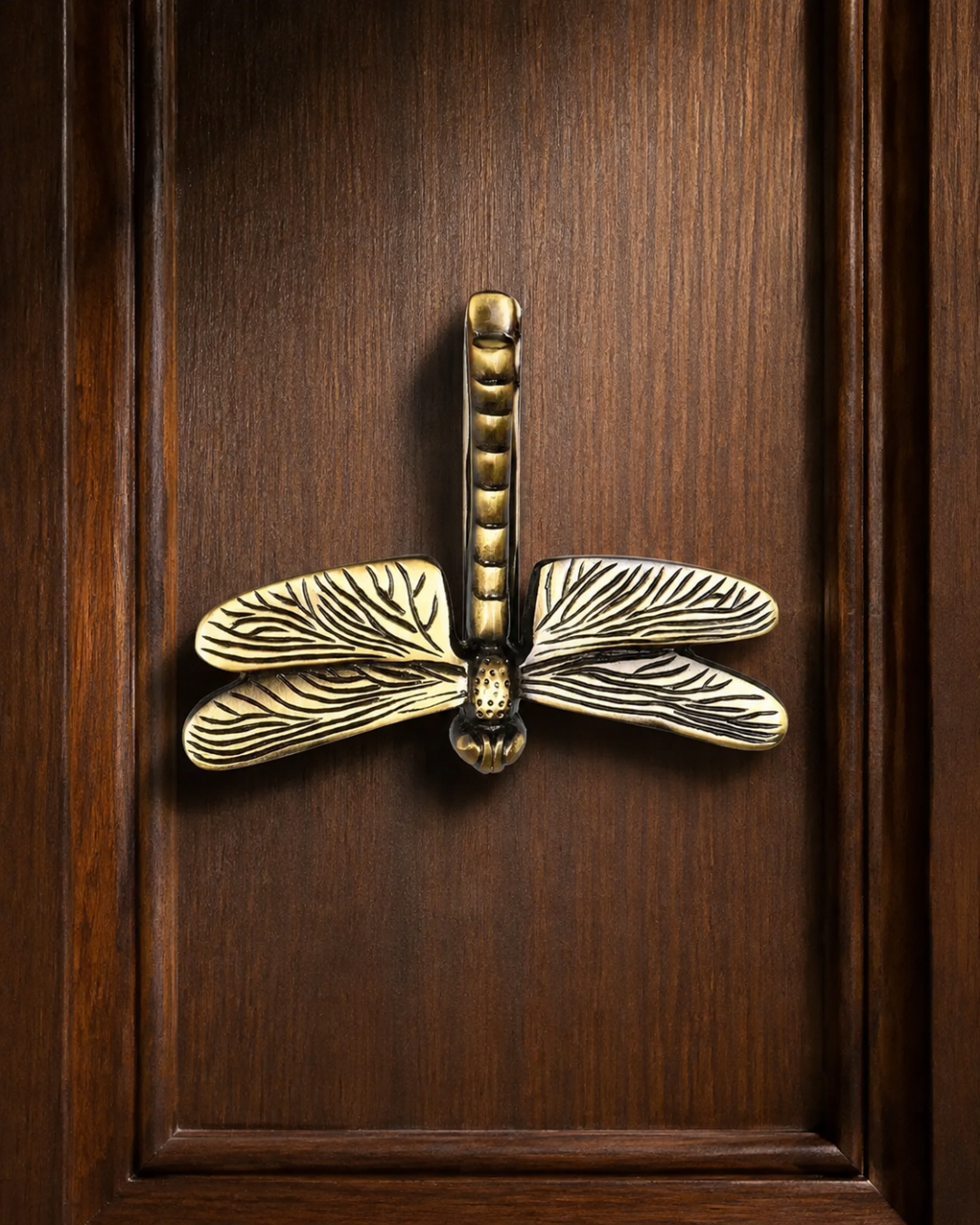 An ornate brass dragonfly door pull handle mounted on a dark brown wooden door.