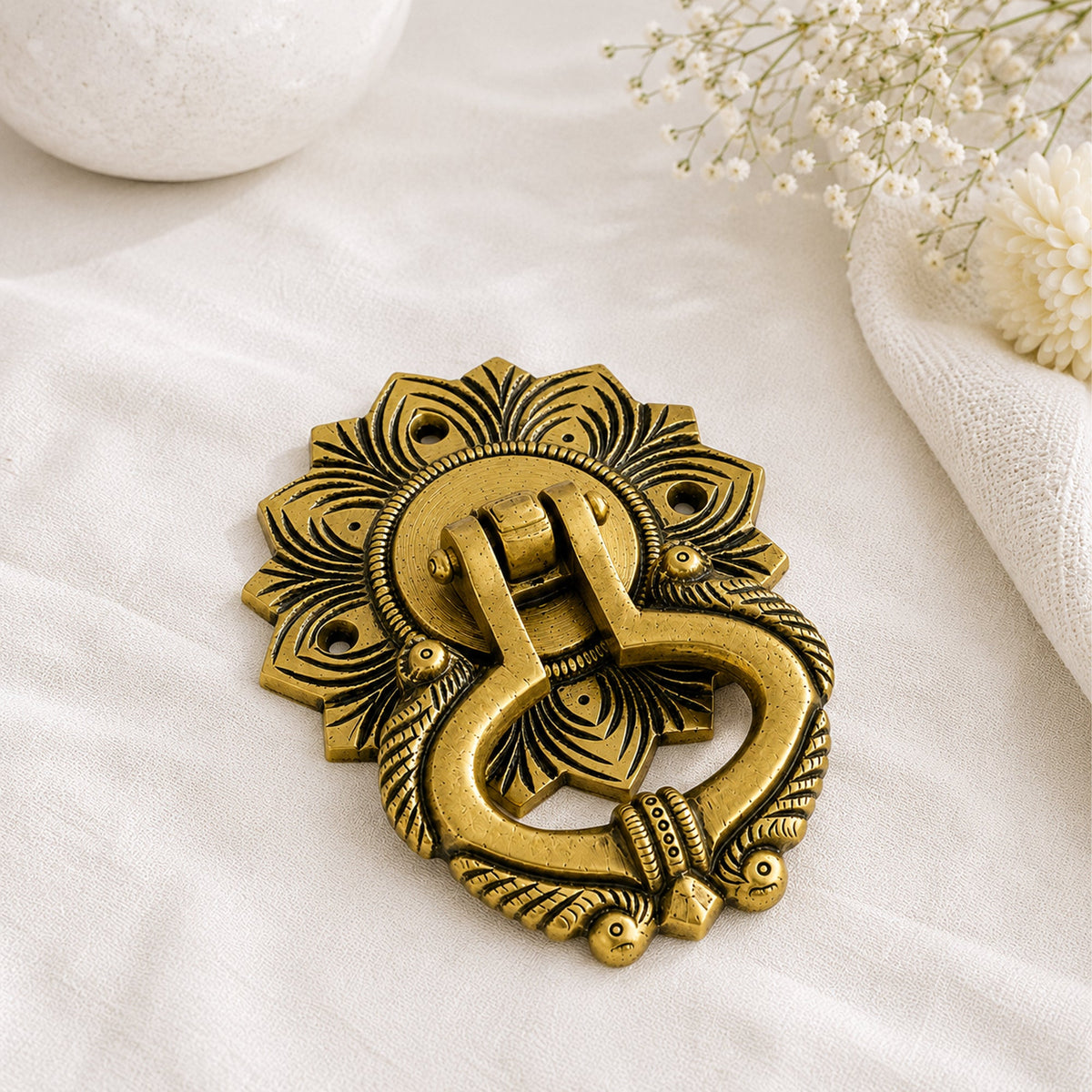 An ornate antique brass door knocker with intricate floral and scrollwork details, displayed on a textured white fabric surface with decorative dried flowers in the corner.