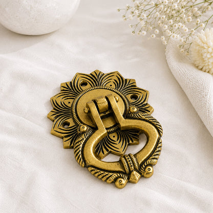 An ornate antique brass door knocker with intricate floral and scrollwork details, displayed on a textured white fabric surface with decorative dried flowers in the corner.