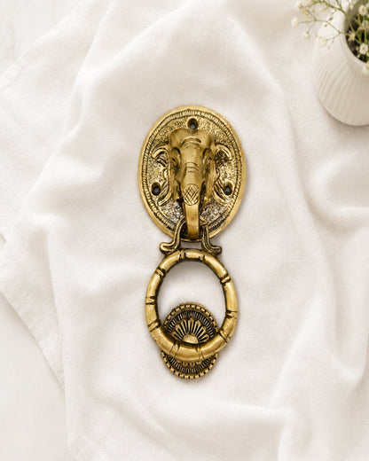 An antique brass elephant head door knocker with a hanging ring, styled on a draped white fabric background with a small white flower pot in the corner.