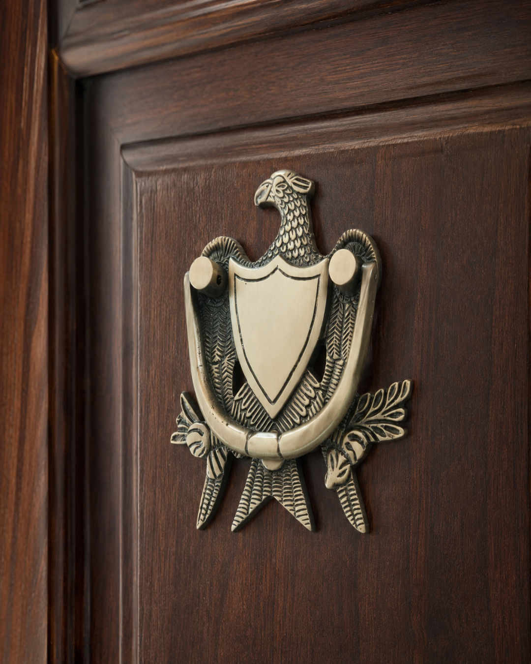 An ornate vintage-style metal door knocker featuring an eagle and shield motif, mounted on a dark brown wooden door.