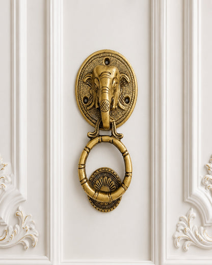 An ornate brass elephant head door knocker with a hanging ring, mounted on a white decorative paneled wall.