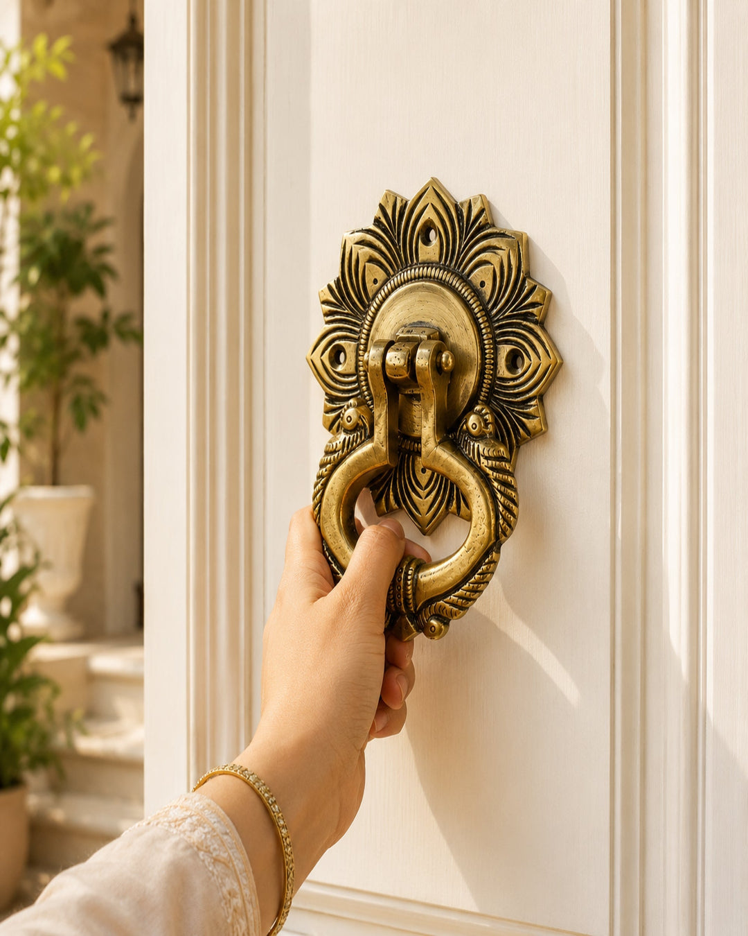 A hand wearing a gold bracelet holds a brass door knocker with an ornate peacock feather design on a white paneled door.
