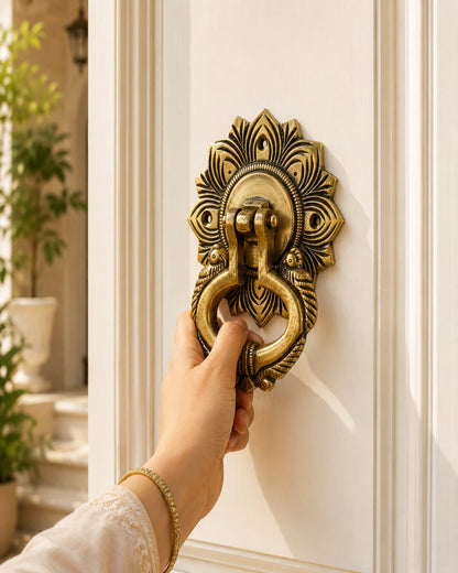 A hand wearing a gold bracelet holds a brass door knocker with an ornate peacock feather design on a white paneled door.